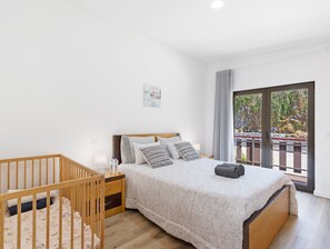 Un dormitorio con una cuna de madera, una cama con ropa de cama estampada, una mesita de noche con una lámpara y vistas a un balcón con barandilla.