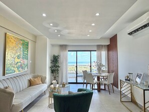 Ein modernes Wohnzimmer mit Sofa, Esstisch, Stühlen, einem grünen Sessel, einem großen abstrakten Gemälde und Meerblick durch große Fenster.