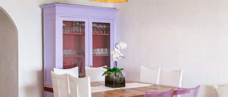 A dining area with a wooden table, white chairs, a purple cabinet with glass doors, and a hanging light fixture.