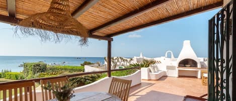 Un patio al aire libre con mesa y sillas, techo de paja y vista al mar.