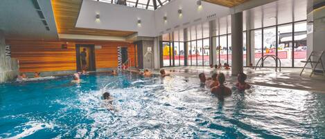 An indoor swimming pool with clear views of the outside through large windows, a wooden ceiling, and visible poolside chairs.
