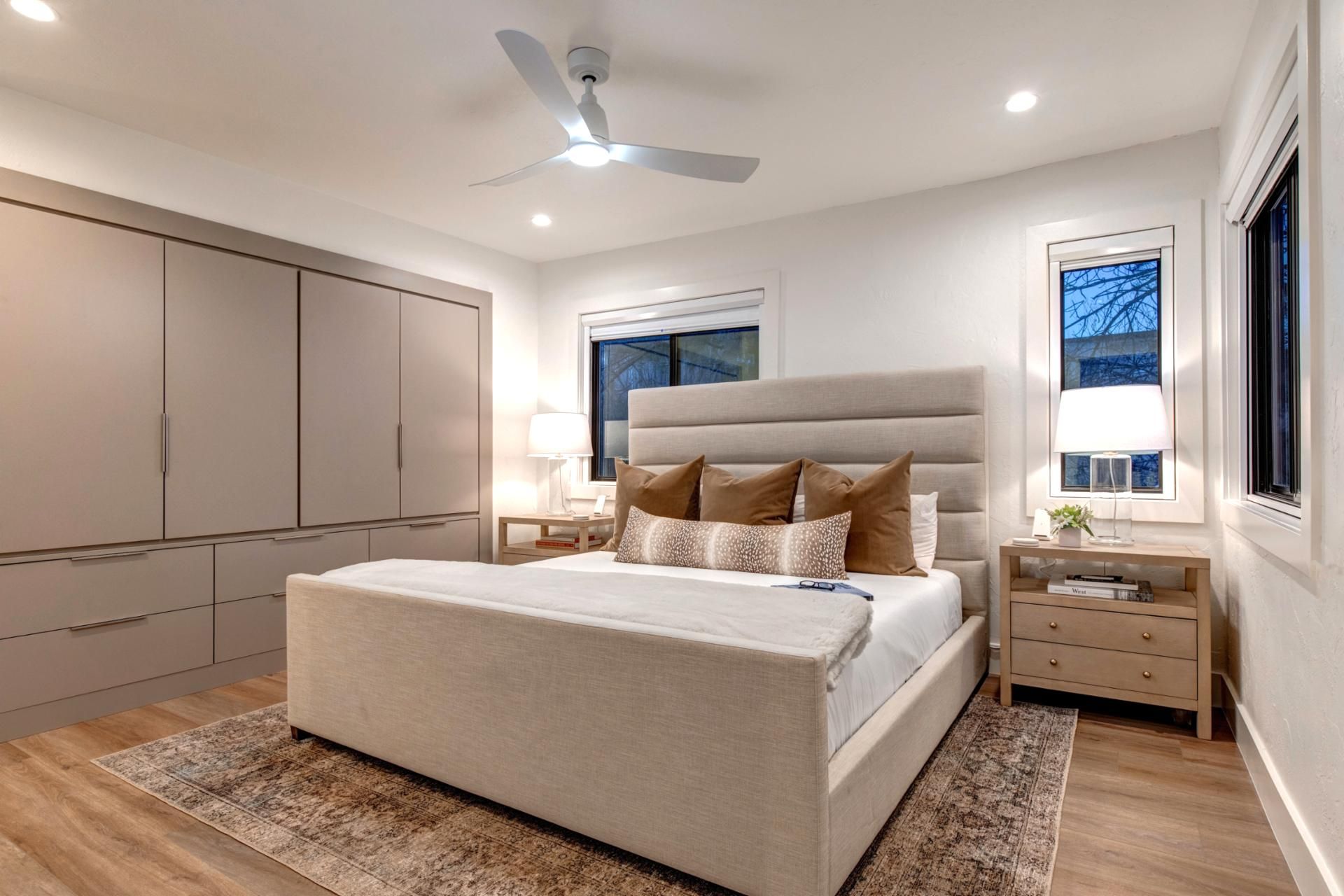 A modern bedroom with a large bed, bedside tables, a ceiling fan, and a window with a view.