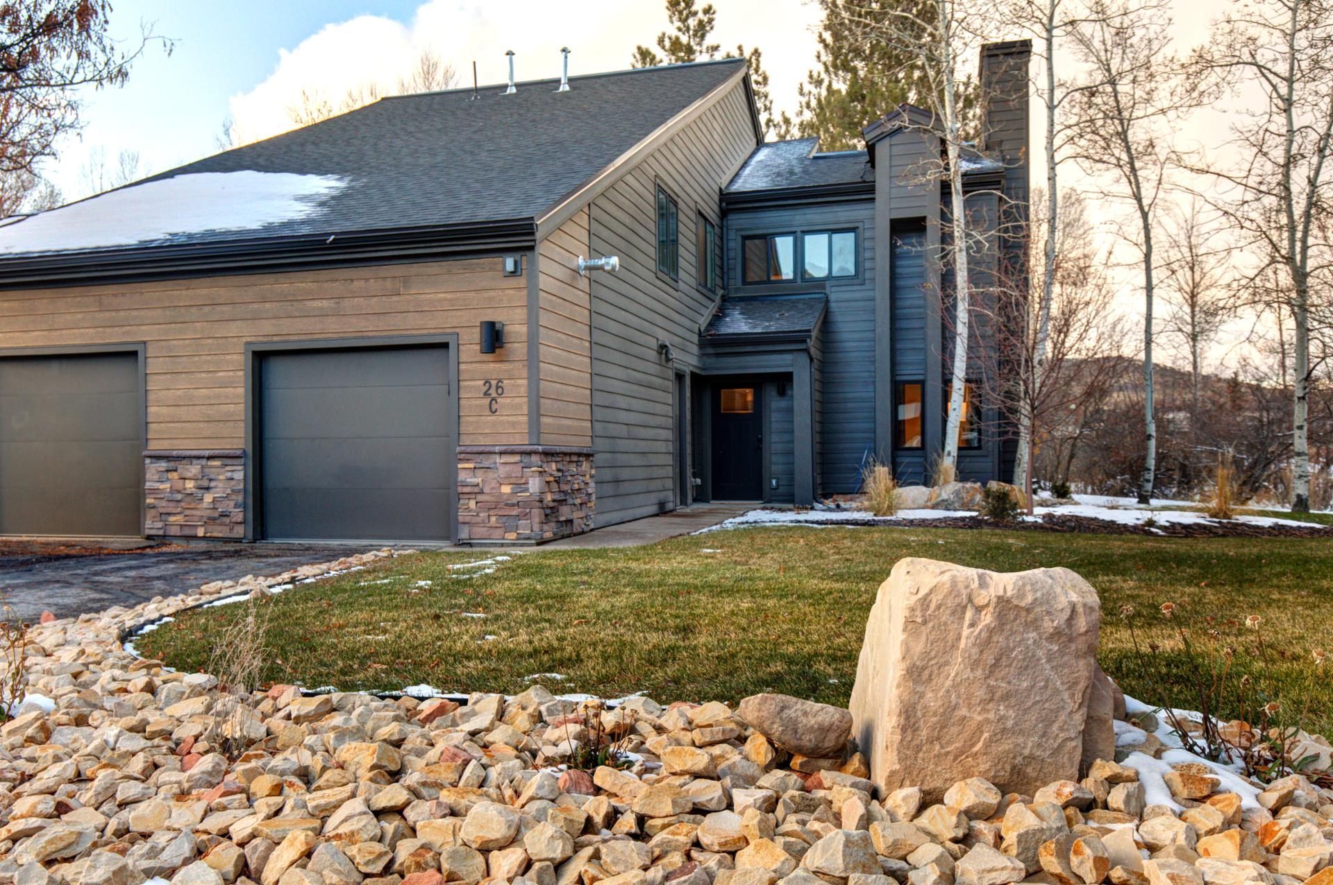 A modern two-story house with a stone foundation, a garage, and a large rock in the front yard.
