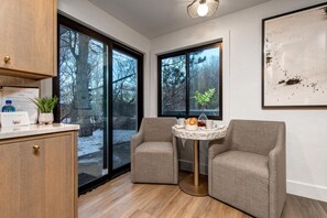 Ein modernes Zimmer mit einem Holzschrank, zwei grauen Sesseln, einem kleinen runden Tisch mit Tablett und großen Fenstern mit Blick auf Bäume und eine verschneite Landschaft.