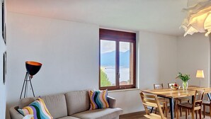 A modern living room with a sofa, a dining table with chairs, a floor lamp, and a window with a view of the sea. - Casa Rivapiana by Interhome (Muralto)