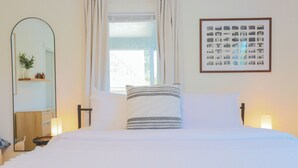 Una cama bien hecha con sábanas blancas, una mesita de noche con una lámpara, un espejo y un cuadro en la pared.