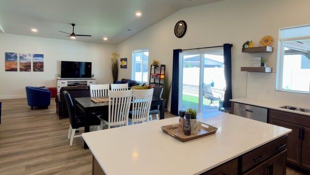 A modern kitchen with a central island, dining area with chairs, a TV mounted on the wall, and a large window with curtains.