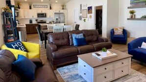 A spacious living area with a brown leather sofa, a blue armchair, and a yellow chair with 'HOME' on it. There is a kitchen in the background with white cabinets and a 'WELCOME' sign.