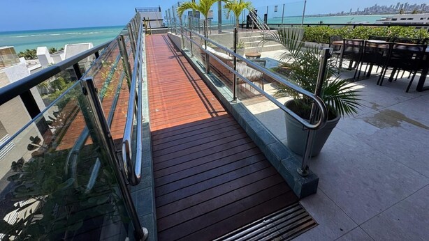 A wooden deck with glass railings leading to a terrace with outdoor seating and a view of the ocean. - Ozean (João Pessoa)