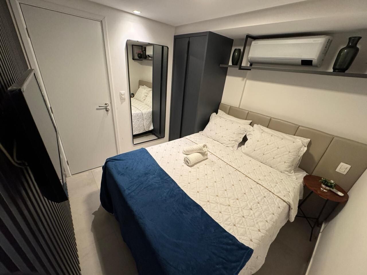 A bedroom with a bed, a wardrobe, a mirror, a shelf with a vase, and a wall-mounted air conditioner.