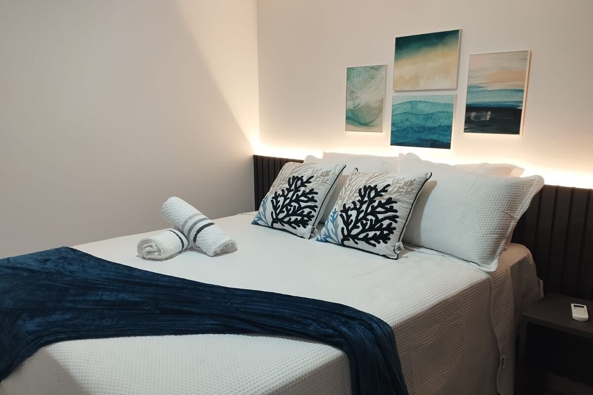Ein ordentlich bezogenes Bett mit blauer Decke, weißen Kissen mit schwarzen Mustern und drei an der Wand hängenden Bildern.