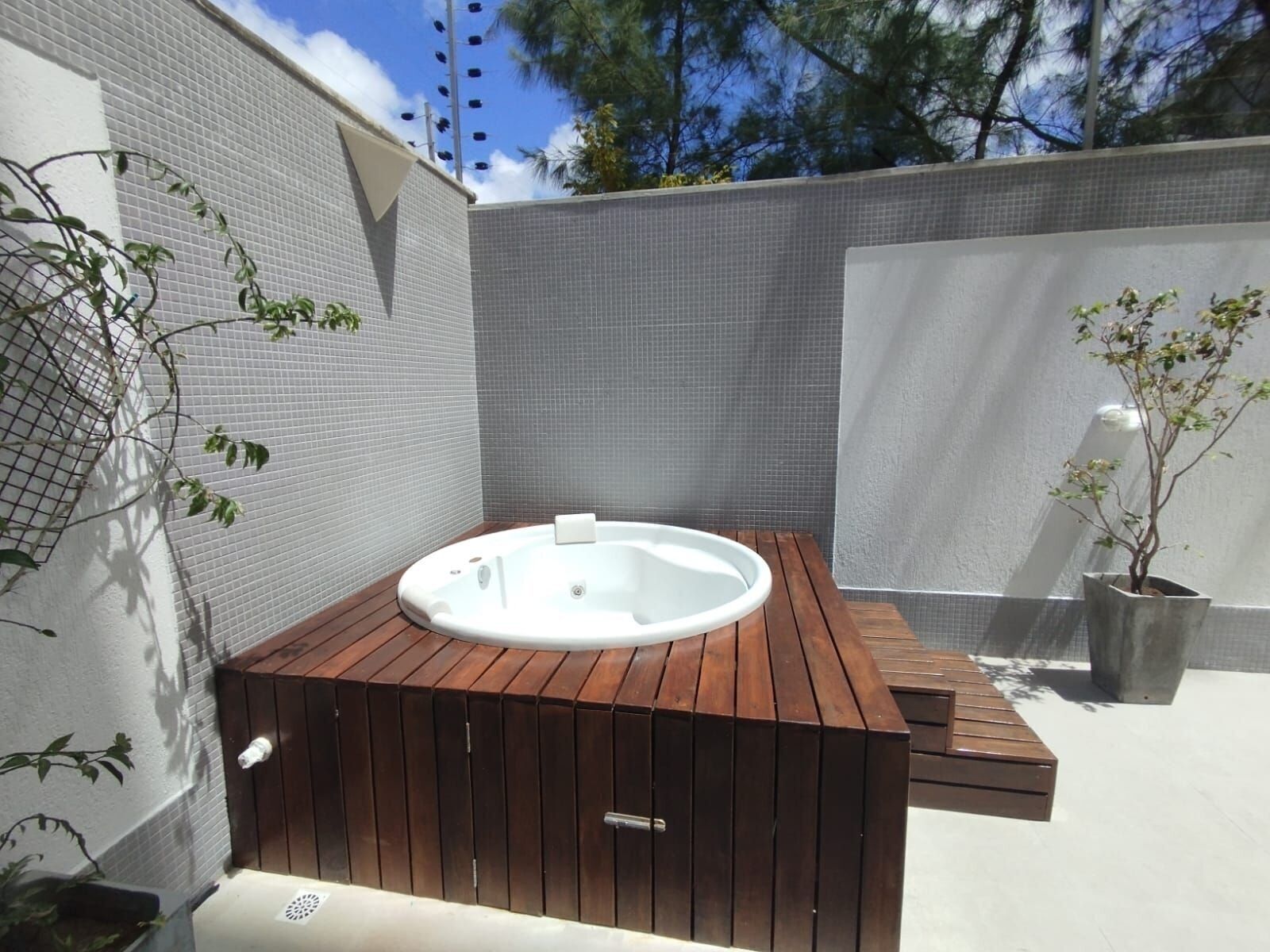 A hot tub in a corner with a wooden deck, surrounded by grey tiled walls and a small potted plant.