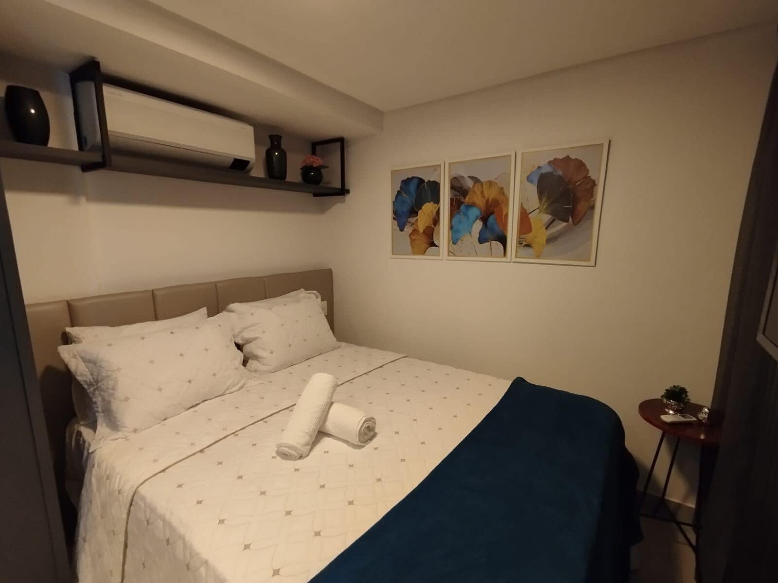 A neatly made bed with a blue blanket and a rolled towel, a bedside table with a plant, and three abstract paintings on the wall.
