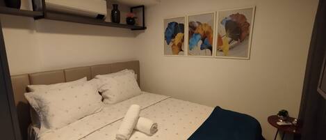 A neatly made bed with a blue blanket and a rolled towel, a bedside table with a plant, and three abstract paintings on the wall.