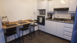 Ett modernt kök med vita skåp, en svart spis, en mikrovågsugn och en kaffebryggare. Det finns två svarta barstolar och ett träbord.