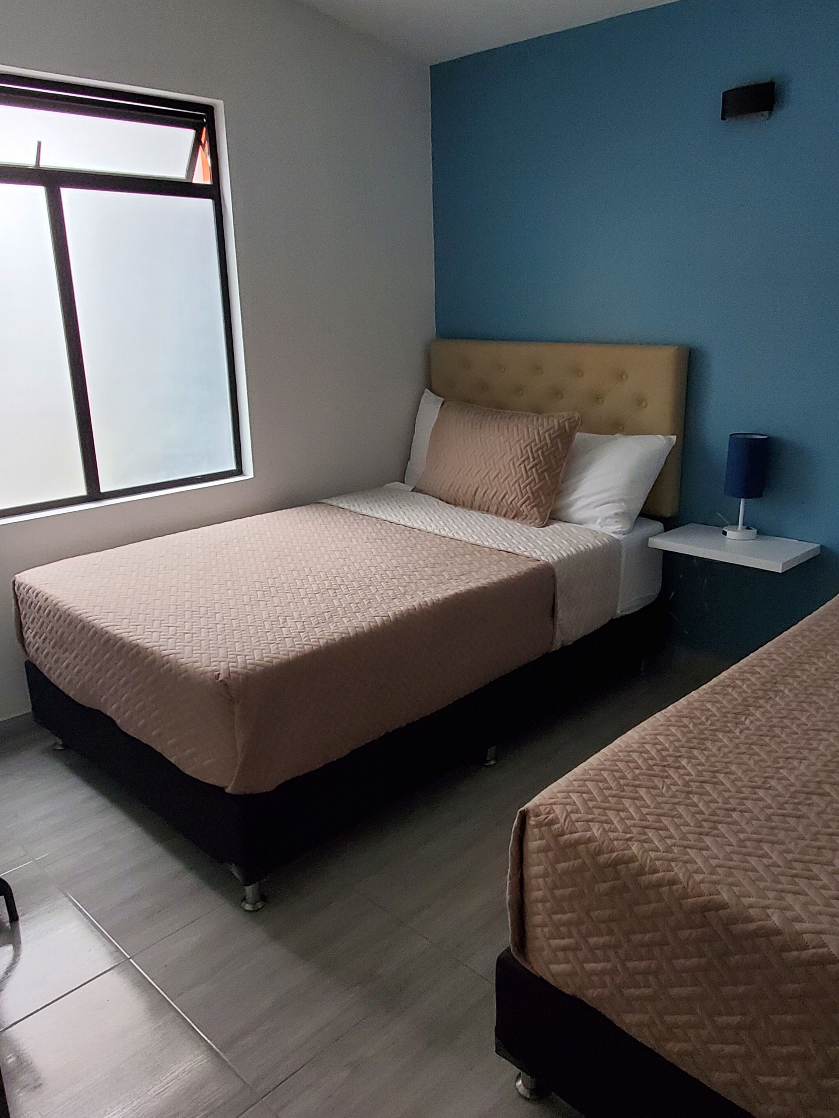 A bedroom with two beds, a window, a nightstand, and a wall-mounted lamp.