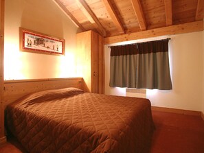 A single bed with a quilted blanket in a room with wooden beams, a window with curtains, and a framed picture on the wall. - Appartment in Vaujany (Les Deux Alpes)