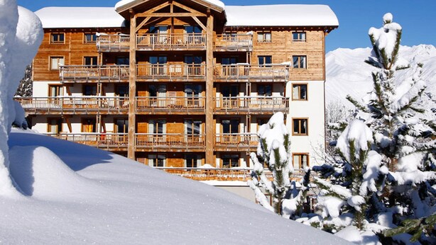 A multi-story wooden hotel with balconies, surrounded by snow-covered trees and mountains. - Appartment in Vaujany (Les Deux Alpes)