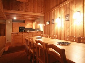 A wooden interior with a dining table and chairs, a kitchen area with wooden cabinets, and a wall-mounted light fixture. - Appartment in Vaujany (Les Deux Alpes)