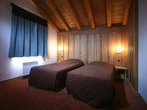 Een kamer met twee bedden, houten balken in het plafond, een raam met gordijnen en wandlampen.