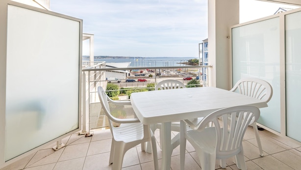 A balcony with a white table and chairs, a view of the sea, and a building in the background. - Ocean-view Duplex for 8 (Plougonvelin)