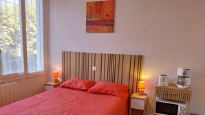 A bed with a red bedspread, two bedside lamps, a wooden headboard, a wall-mounted artwork, a small shelf with a coffee maker, a microwave, and a trash bin. - Apartment “Sauges 16” with Wi-Fi (Cazaubon)