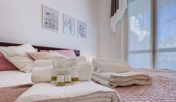 A neatly made bed with folded towels, a bedside table with bottles, and framed artwork on the wall.