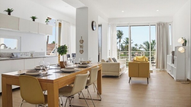 Une cuisine moderne avec des meubles blancs, un coin repas avec une table et des chaises en bois, un canapé et une grande fenêtre donnant sur des palmiers.