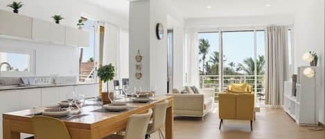 A modern kitchen with white cabinets, a dining area with a wooden table and chairs, a sofa, and a large window with a view of palm trees.