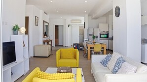 Un salon moderne avec un canapé jaune, un divan blanc et un téléviseur. On y trouve également un coin repas avec une table et des chaises, et une cuisine à l’arrière-plan.