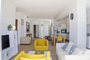 A modern living room with a yellow sofa, a white couch, and a TV. There is a dining area with a table and chairs, and a kitchen in the background. - Your very own oasis awaits you! (Bavaro)