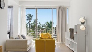 Un salon moderne avec un canapé, un pouf et un meuble pour la télévision, de grandes fenêtres avec des rideaux et un balcon agrémenté de palmiers.