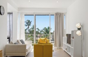 A modern living room with a sofa, ottoman, and TV stand, large windows with curtains, and a balcony with palm trees. - Your very own oasis awaits you! (Bavaro)