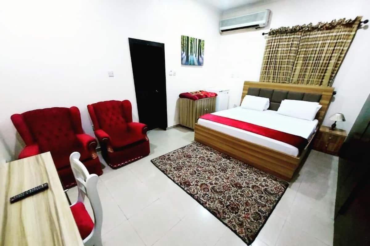 Une chambre d’hôtel avec un lit, deux fauteuils rouges, une table en bois, un tapis à motifs, une porte, un climatiseur et un tableau accroché au mur.