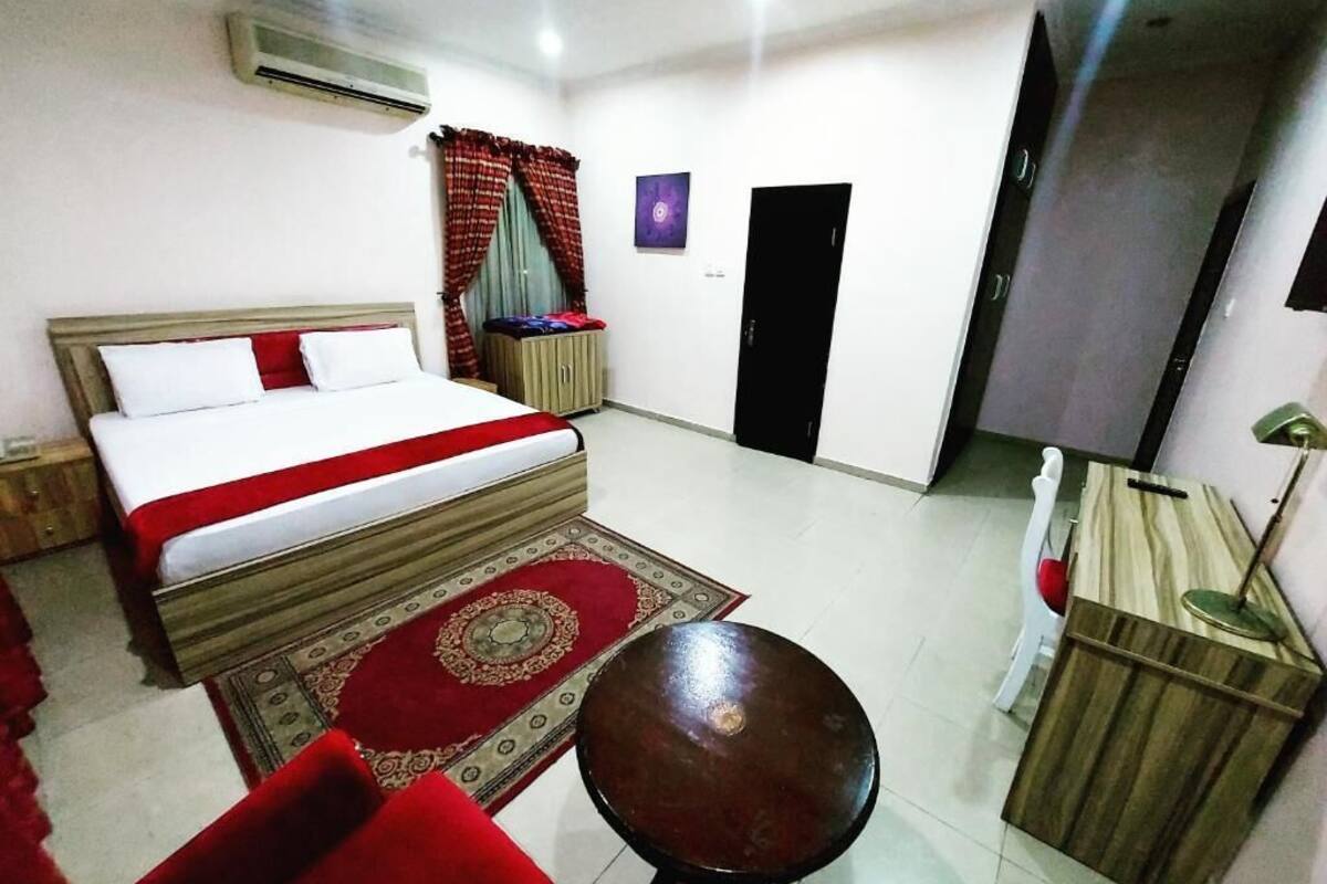Une chambre d’hôtel comprenant un lit, une petite table ronde, une armoire, une chaise rouge, un tapis aux motifs rouges et dorés, un climatiseur fixé au mur, une porte et un luminaire fixé au mur.