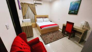 A bedroom with a bed, a desk, a chair, a red sofa, and a painting on the wall.