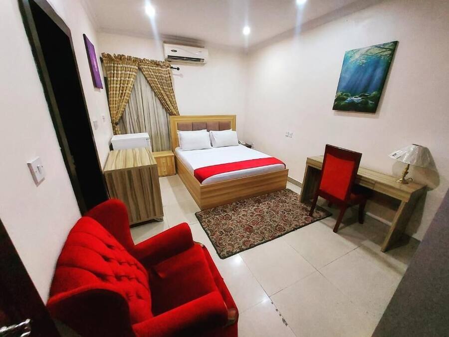A bedroom with a bed, a desk, a chair, a red sofa, and a painting on the wall.