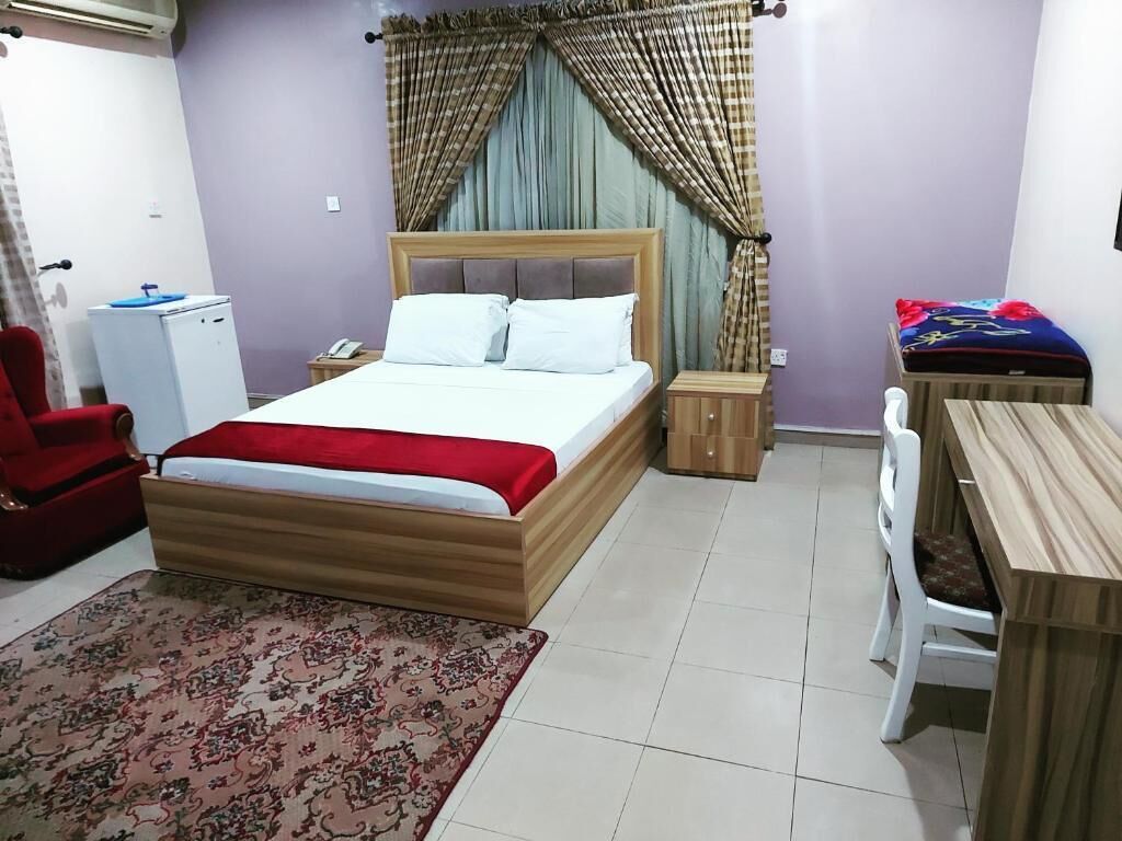 A bedroom with a bed, a desk, a chair, a red armchair, a small table, a refrigerator, and curtains.