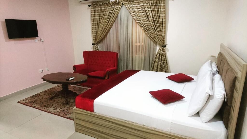Un dormitorio con cama, un sillón rojo, una mesita y un televisor fijado a la pared.