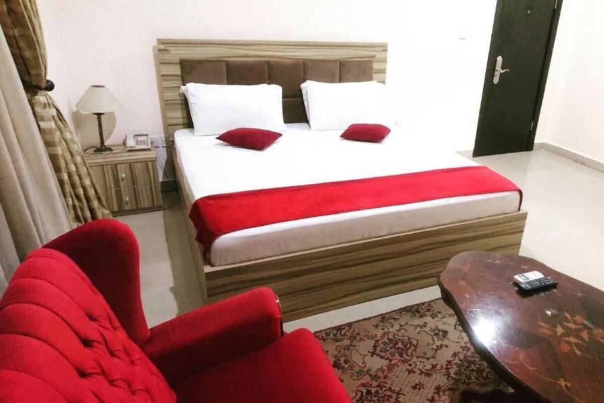 Une chambre d’hôtel avec un lit, un fauteuil rouge, une table d’appoint en bois et une porte.