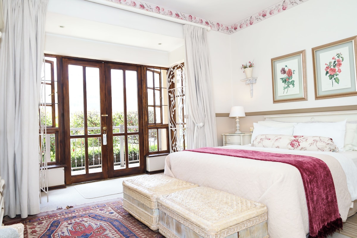 A bedroom with a large window, a bed with white linens, a bedside table, and a wall-mounted lamp.