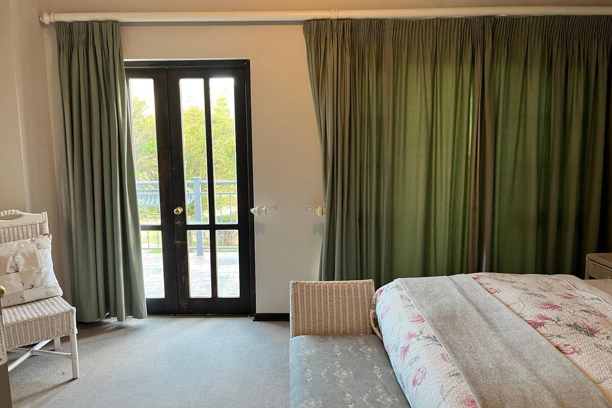 Habitación de hotel con cama, una silla, una mesita, ventana con cortinas y puerta que da al exterior.