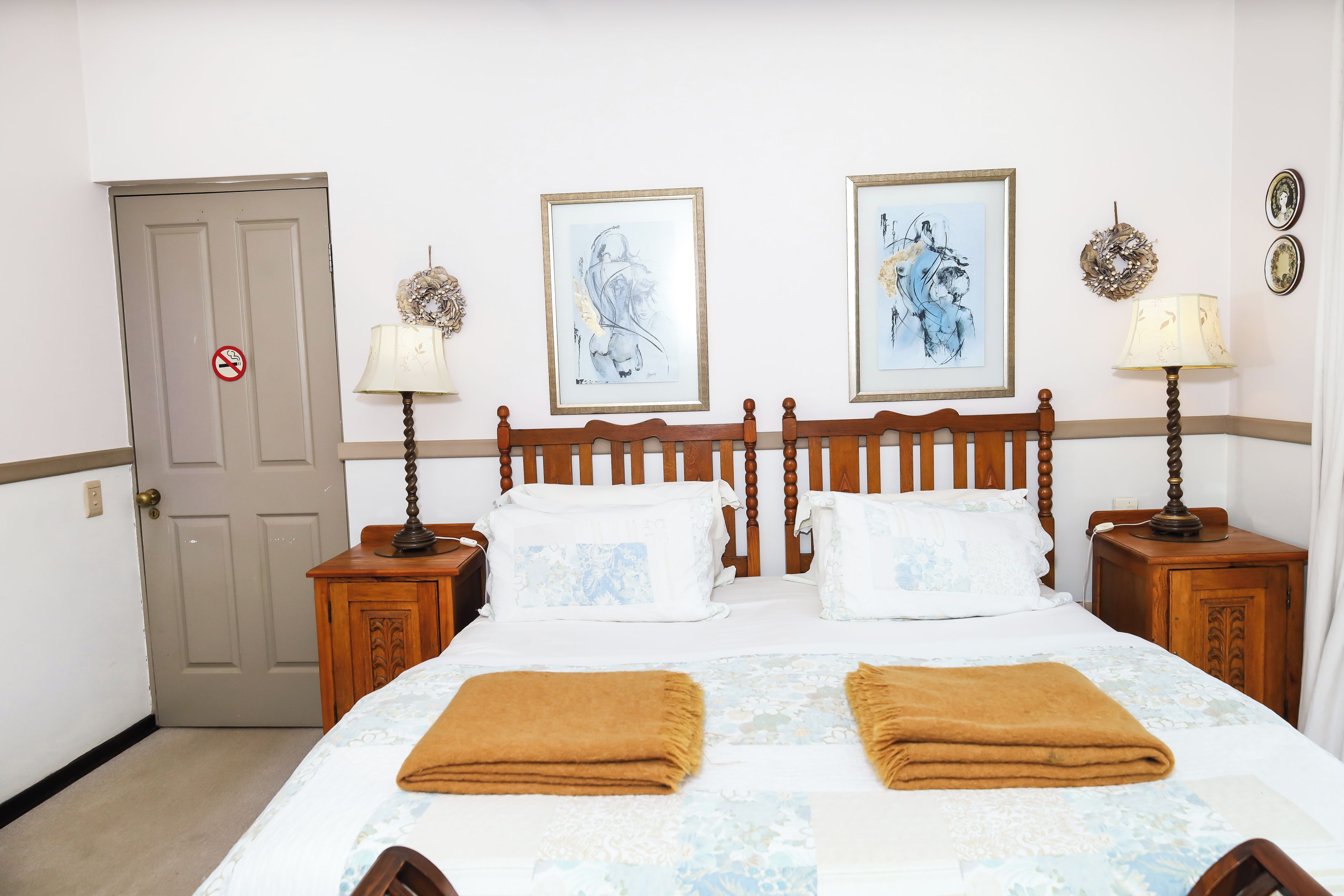 A hotel room with a wooden bed, two nightstands, a door with a no-smoking sign, and two framed artworks on the wall.