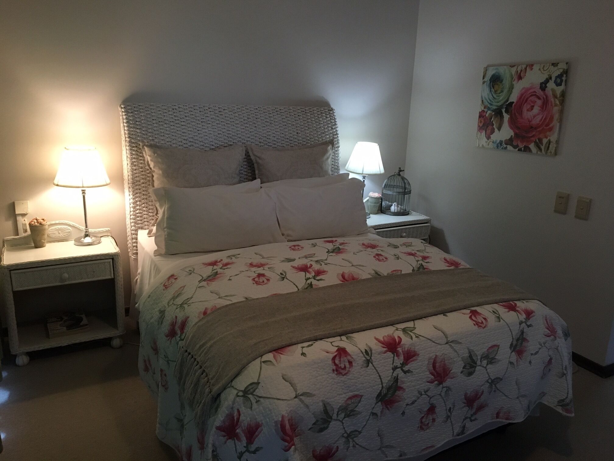 A neatly made bed with floral bedding, two bedside lamps, a nightstand with a lamp, and a wall-mounted floral artwork.