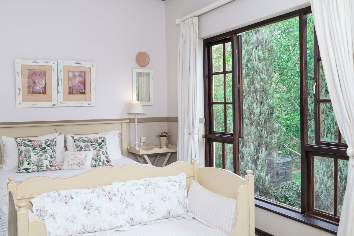 A bedroom with a bed, a bedside table, a lamp, a window with curtains, and framed pictures on the wall.