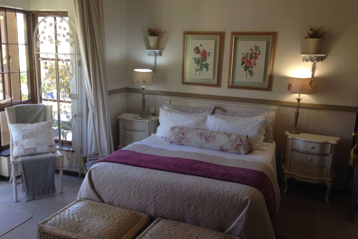 Un dormitorio con cama, dos mesitas de noche, una silla, una ventana con cortinas y lámparas de pared.
