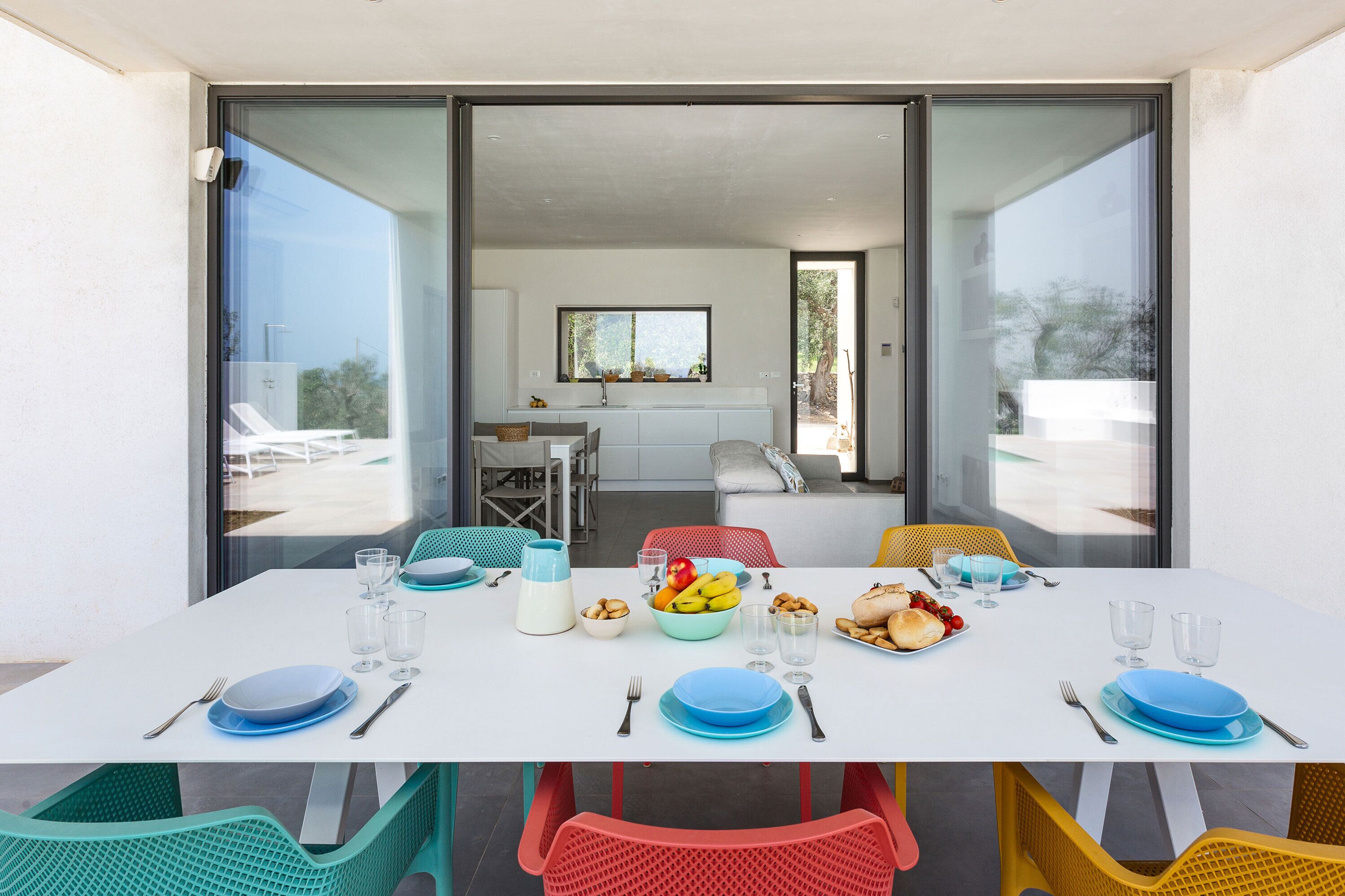 Mesa lista con platos, vasos y cubiertos, con vistas a una cocina y una zona de estar al aire libre.
