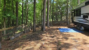 A forested campsite with a RV, a wooden deck, and a blue mat.