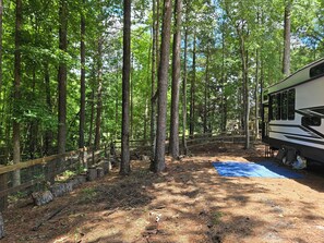 A forested campsite with a RV, a wooden deck, and a blue mat.