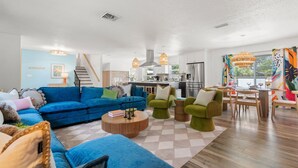 Ein modernes Wohnzimmer mit einem blauen Sofa, grünen Sesseln, einem Holztisch und einer Küche im Hintergrund.